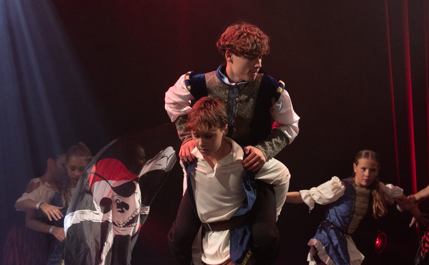 One boy dancer piggybacking another with girls in background on stage pirate routine