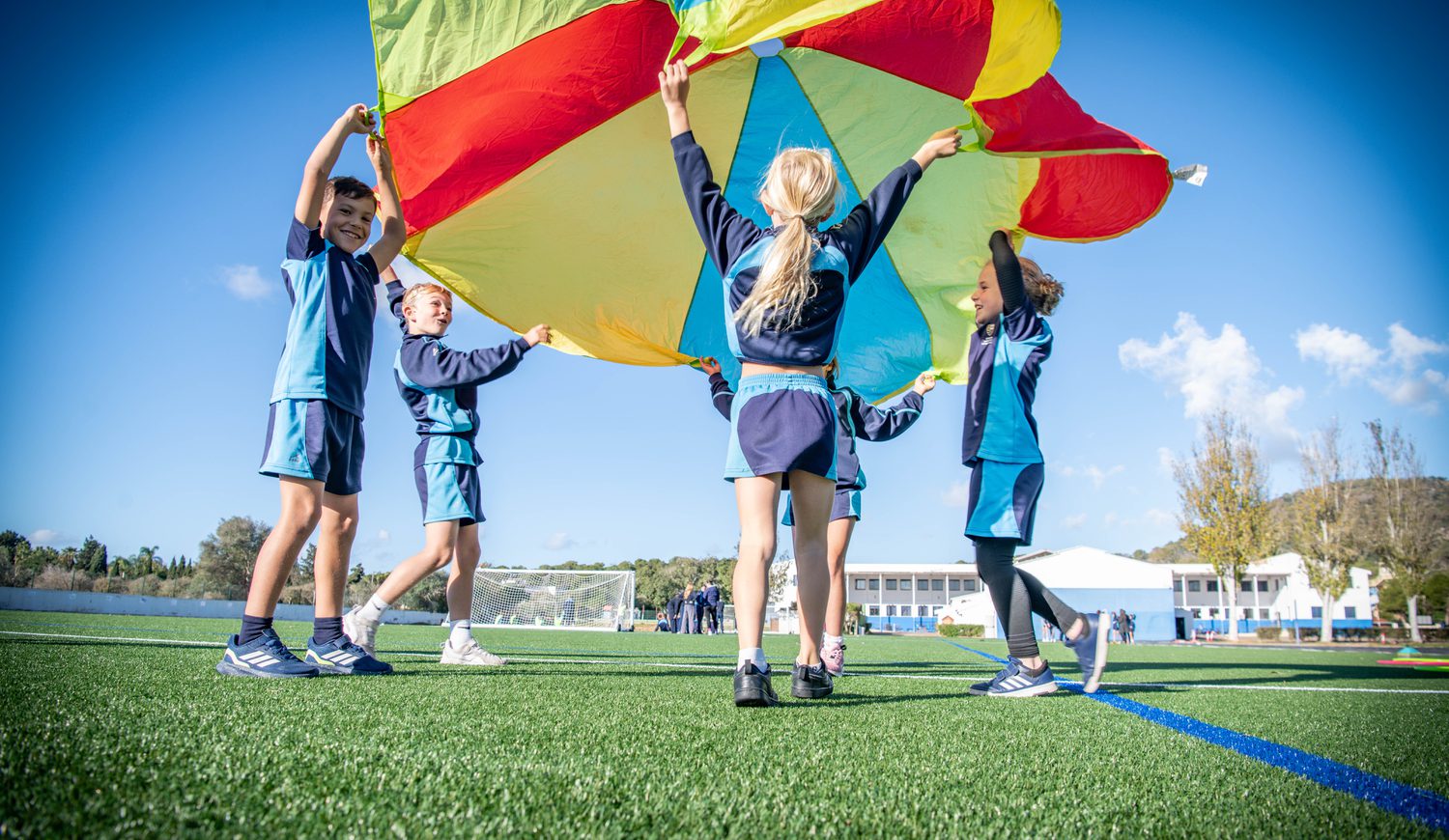 Five primary kids holding up a colour parachute on the Astro Turf with school in background