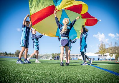 Five primary kids holding up a colour parachute on the Astro Turf with school in background