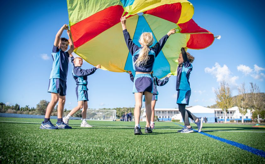 Five primary kids holding up a colour parachute on the Astro Turf with school in background