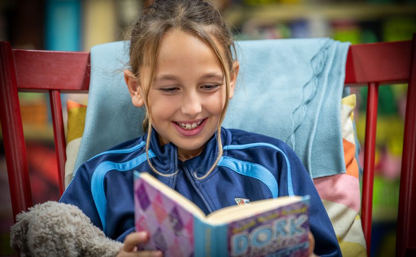 Primary girl reading a book on a chair in the library with a fluffy toy in arm