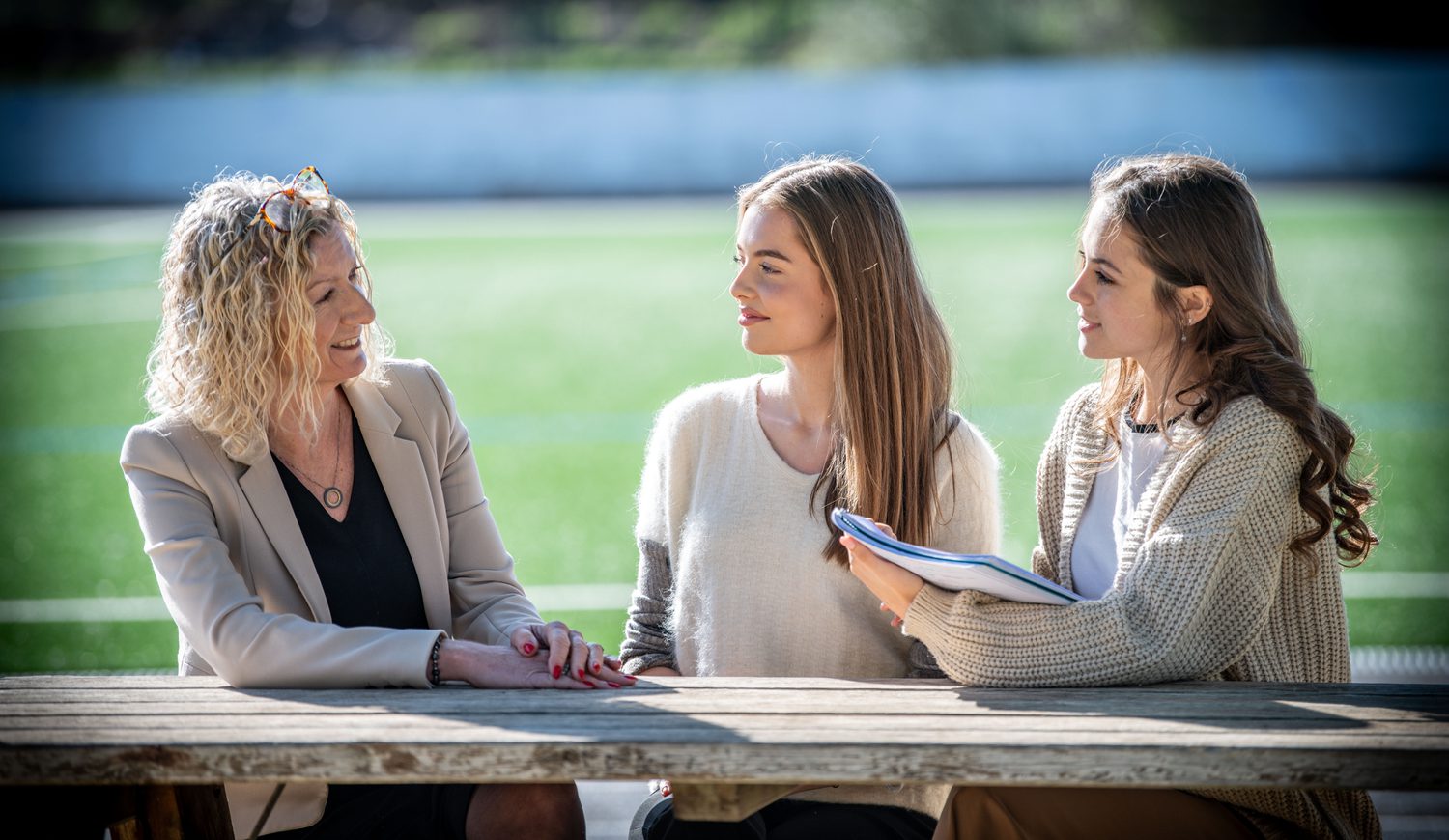 Principal chatting to two Sixth Form girls outside on a bench with Astro Turf in background