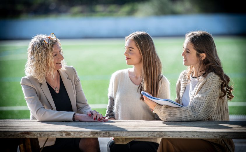 Principal chatting to two Sixth Form girls outside on a bench with Astro Turf in background