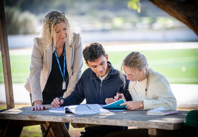 Principal leaning over looking at two Sixth Form students working outside on a bench