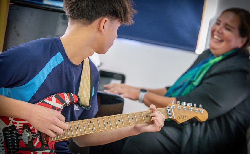 Secondary boy playing guitar looking at his music teacher on the piano