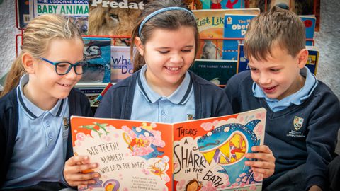 Three Primary children smiling while reading a book in the reading corner