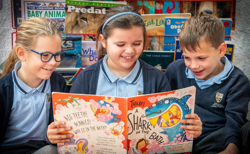 Three Primary children smiling while reading a book in the reading corner