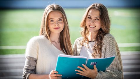 Two Sixth Form girls smiling at the camera with a book they are sharing