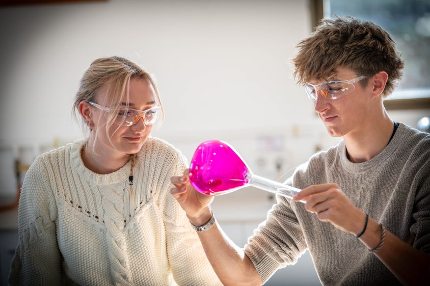 Two Sixth Form students looking at a pink glass beaker in the science lab with glasses on