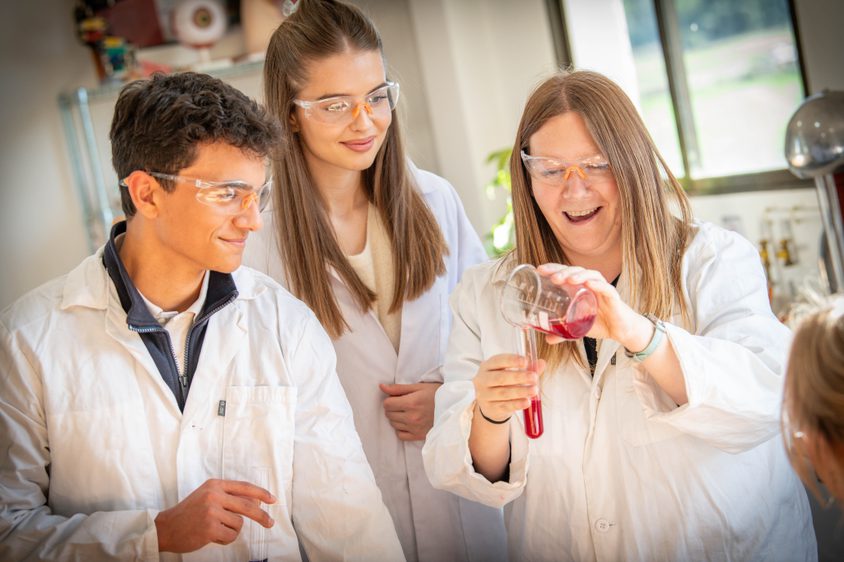 Two Sixth Form students watching a teacher pouring liquid into a tube in the science lab