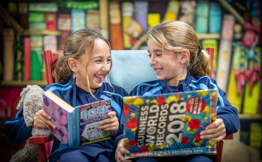 Two Year 5 girls laughing while reading books on a chair in the library