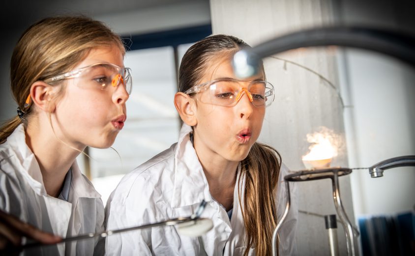Two secondary girls in lab coats and glasses blowing onto a petri dish with flames